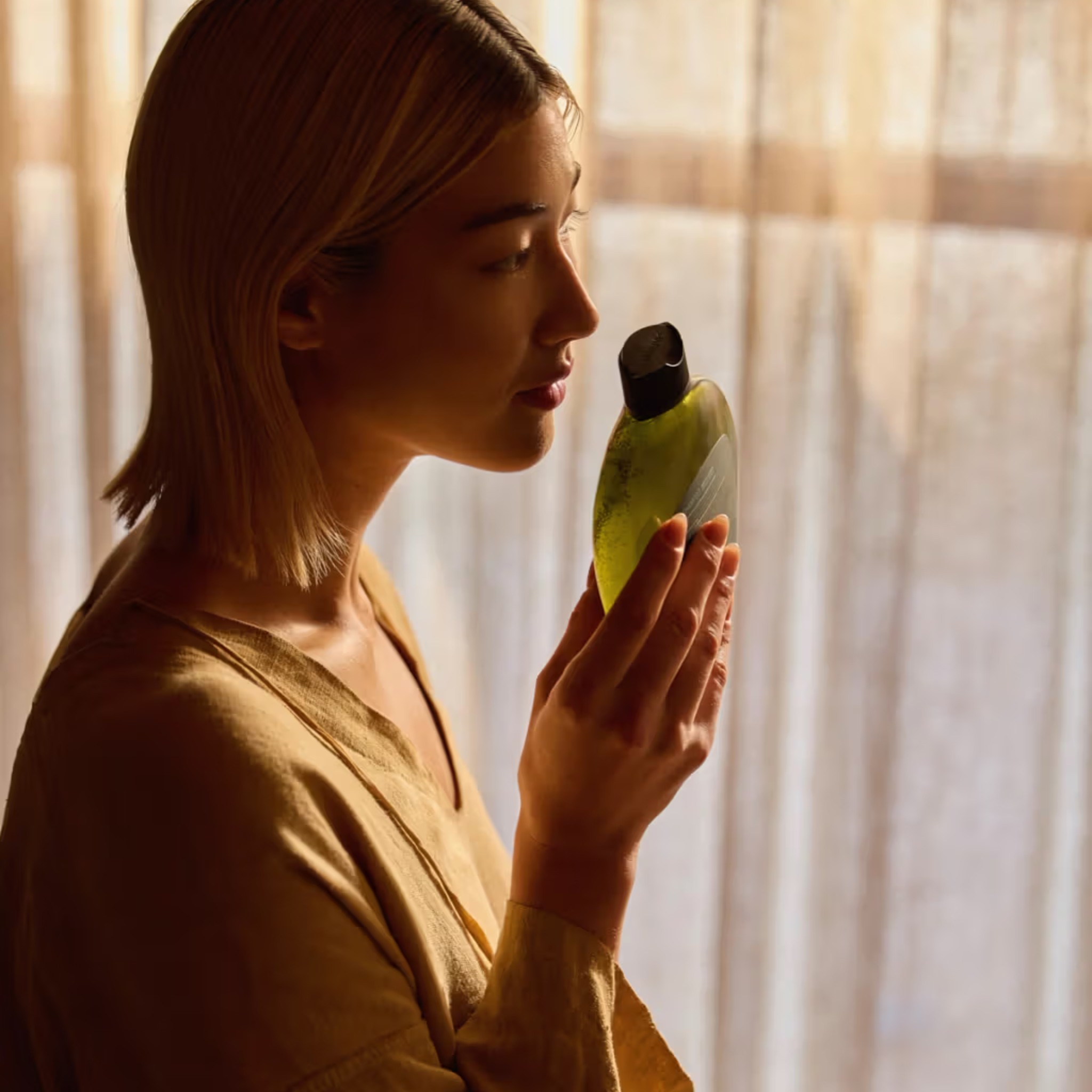Woman holding a green bottle of Antipodes Peanch Freesia Shampoo with a black cap in front of a softly lit curtain.