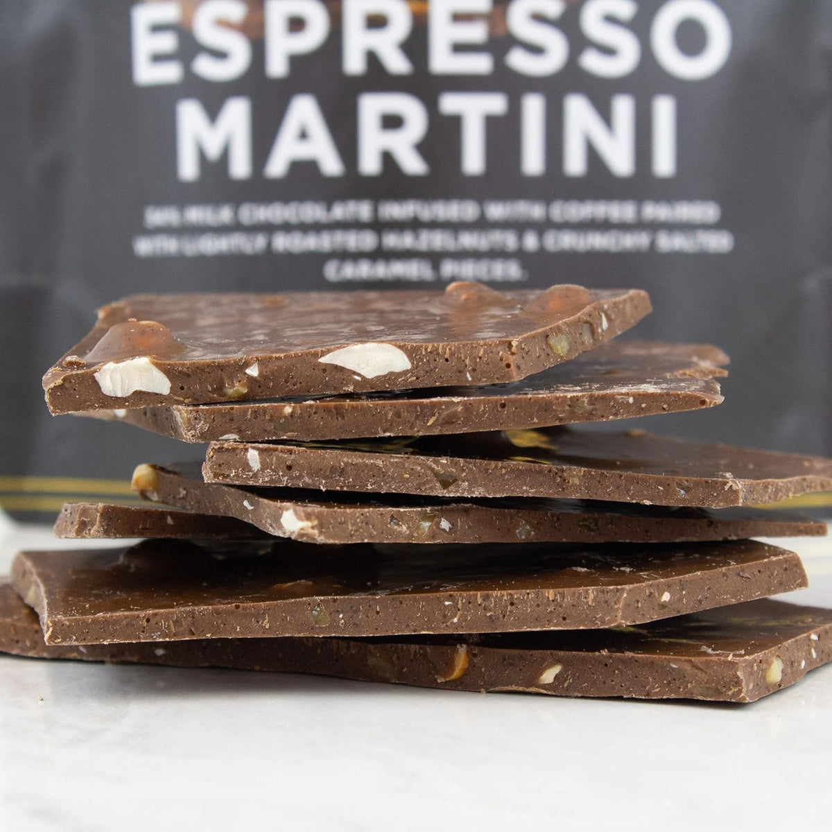 A stack of dark chocolate bark pieces with visible nuts and caramel pieces, set against a dark bag labeled 'ESPRESSO MARTINI'.