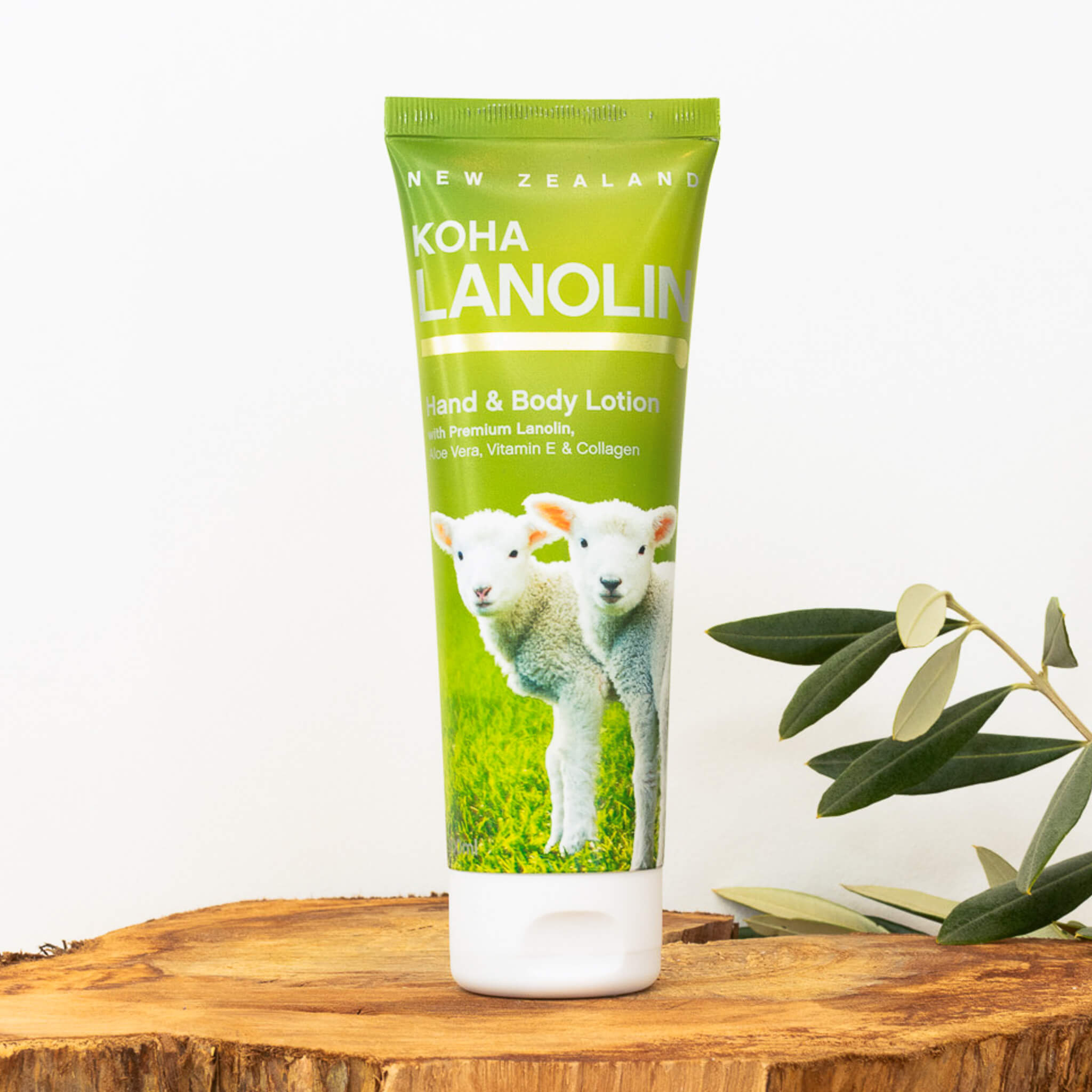 A tube of Koha Lanolin Hand & Body Lotion with a green label featuring images of sheep on a wooden surface with a plant in the background.