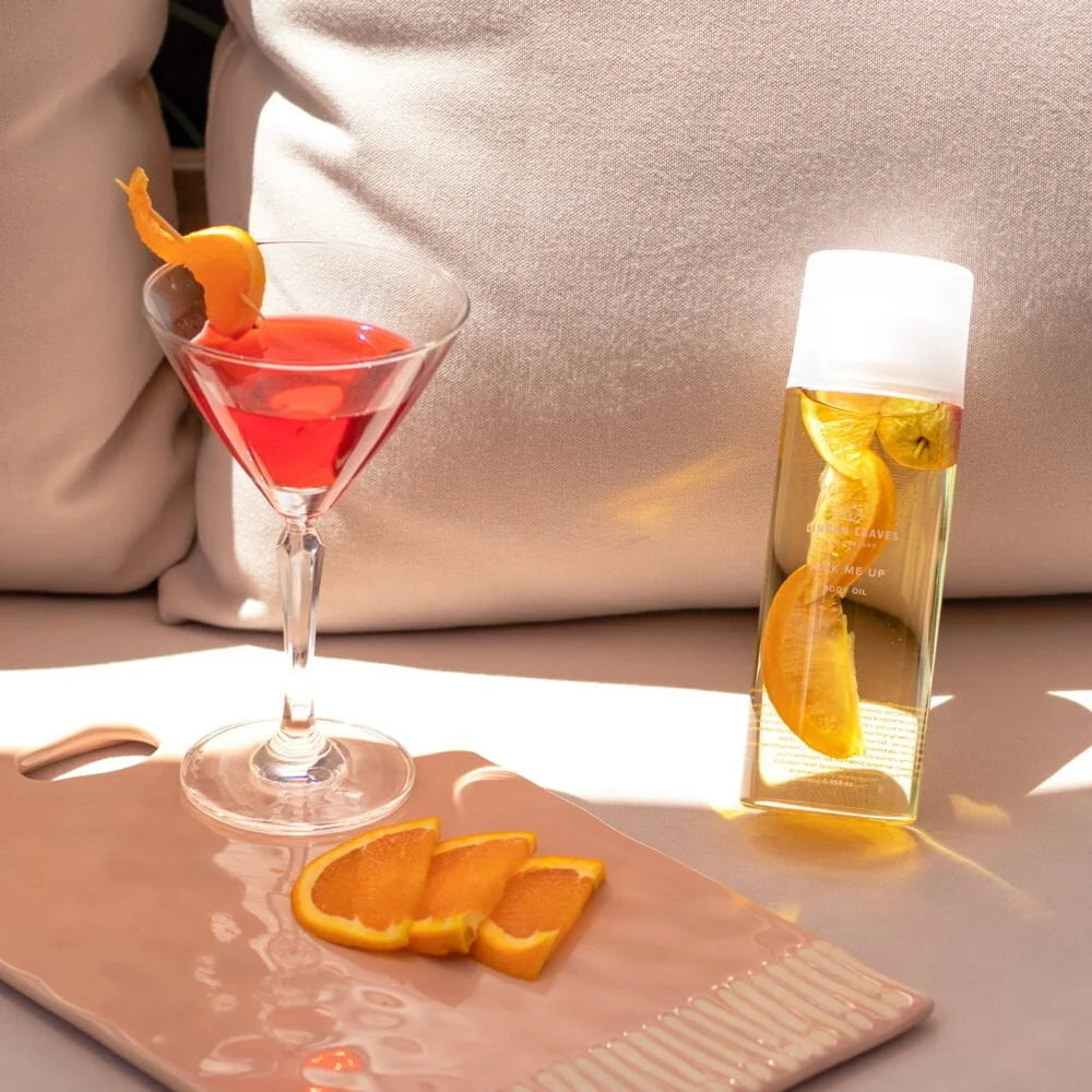 Cocktail with an orange twist and a bottle of Linden Leave memories body oil ml on a table.