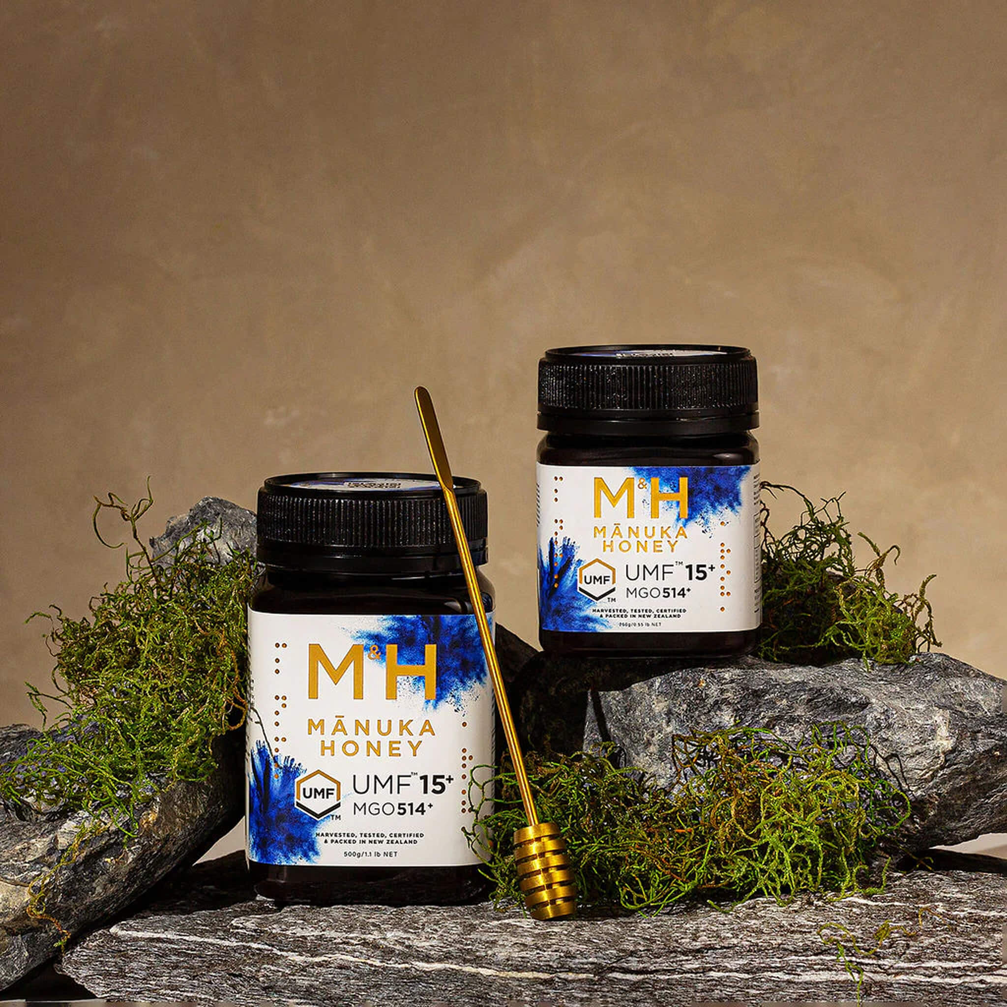 Two jars of M&H Manuka Honey UMF 15+ 250g and 500g with a honey dipper on a natural stone background.