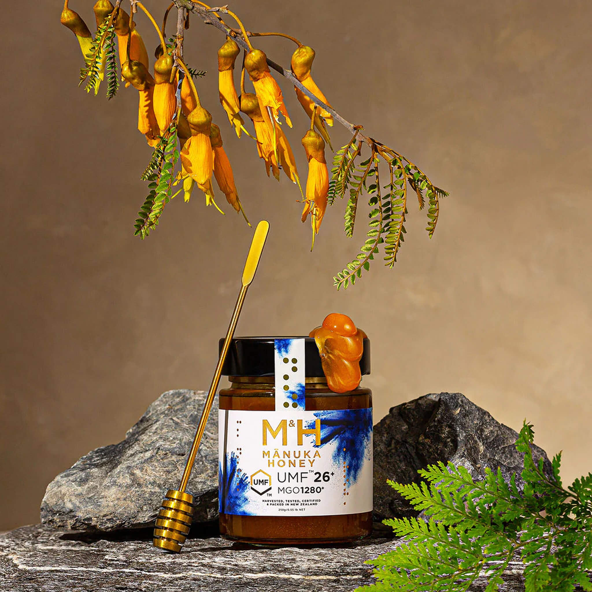 M&H Manuka honey UMF26+ jar with a dipper on rocks, surrounded by flowers and plants.