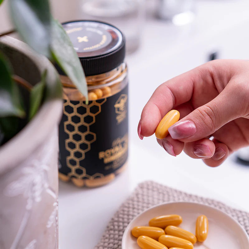 Hand holding a yellow Manuka South Royal Jelly extra strength supplement capsule with a bottle and more capsules in the background.