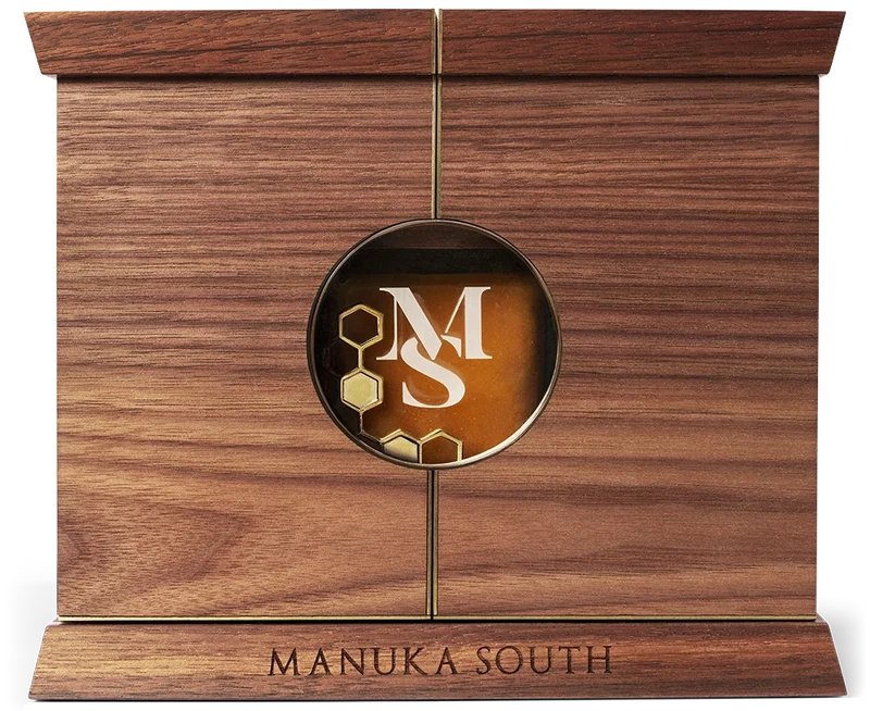 A rectangular wooden plaque or keepsake box with a warm medium-brown wood grain finish, featuring a circular brass emblem in the center with a white stylized 'MS' monogram and gold honeycomb details, and the words 'MANUKA SOUTH' engraved on the base. Manuka South Manuka Hosney UMF35+on a workshop bench