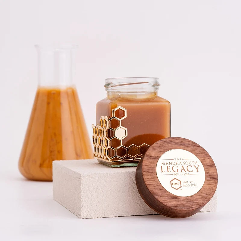 A jar candle with amber-brown wax sits on a light block, with its round wooden lid placed in front. The jar has a gold honeycomb pattern around the lower half, and the lid is printed with the brand name 'Longevity by Manuk & South'. Manuka South Manuka Honey UMF35+.