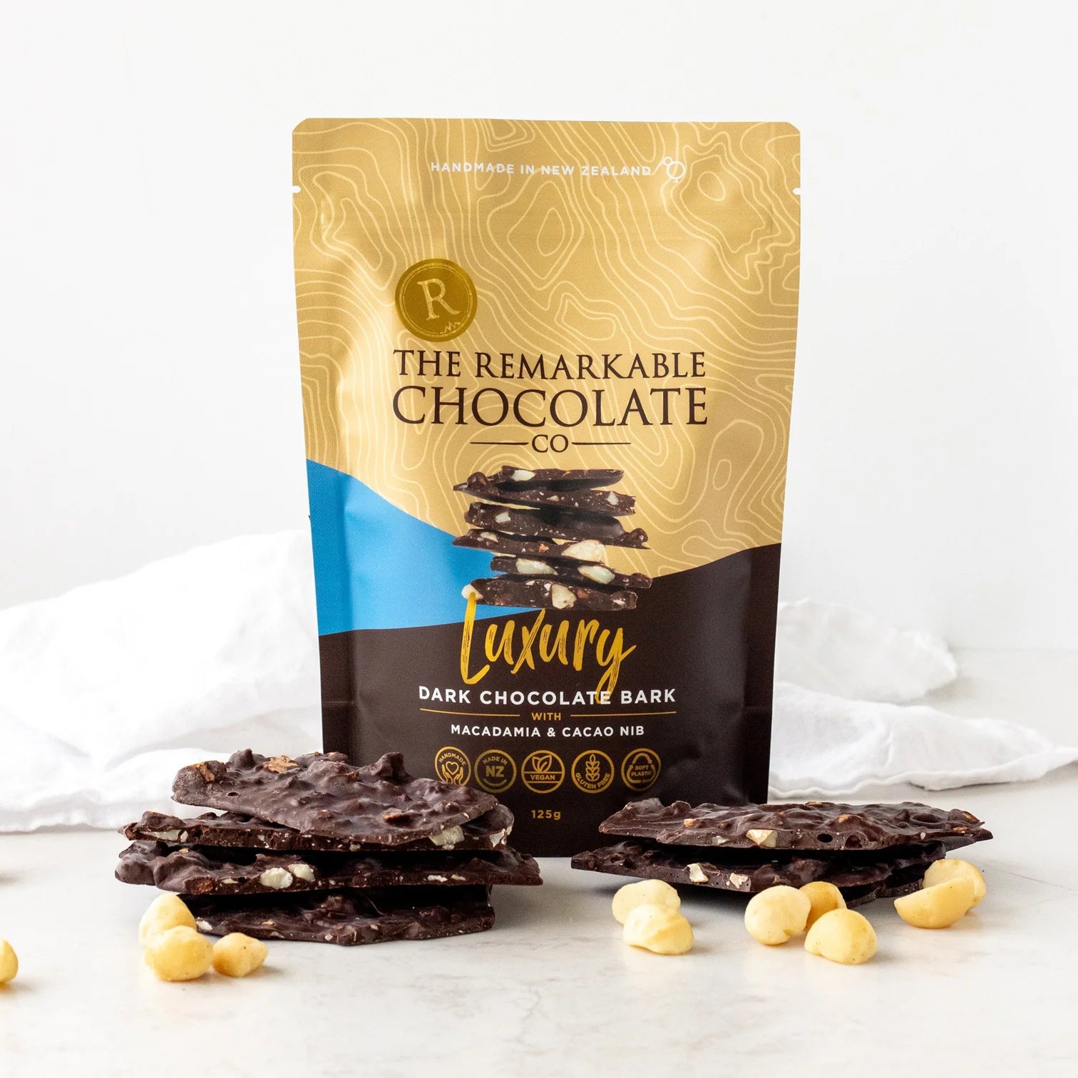 Packaging of The Remarkable Chocolate Co. Luxury Dark Chocolate Bark with chocolate pieces and macadamia nuts on a white background.