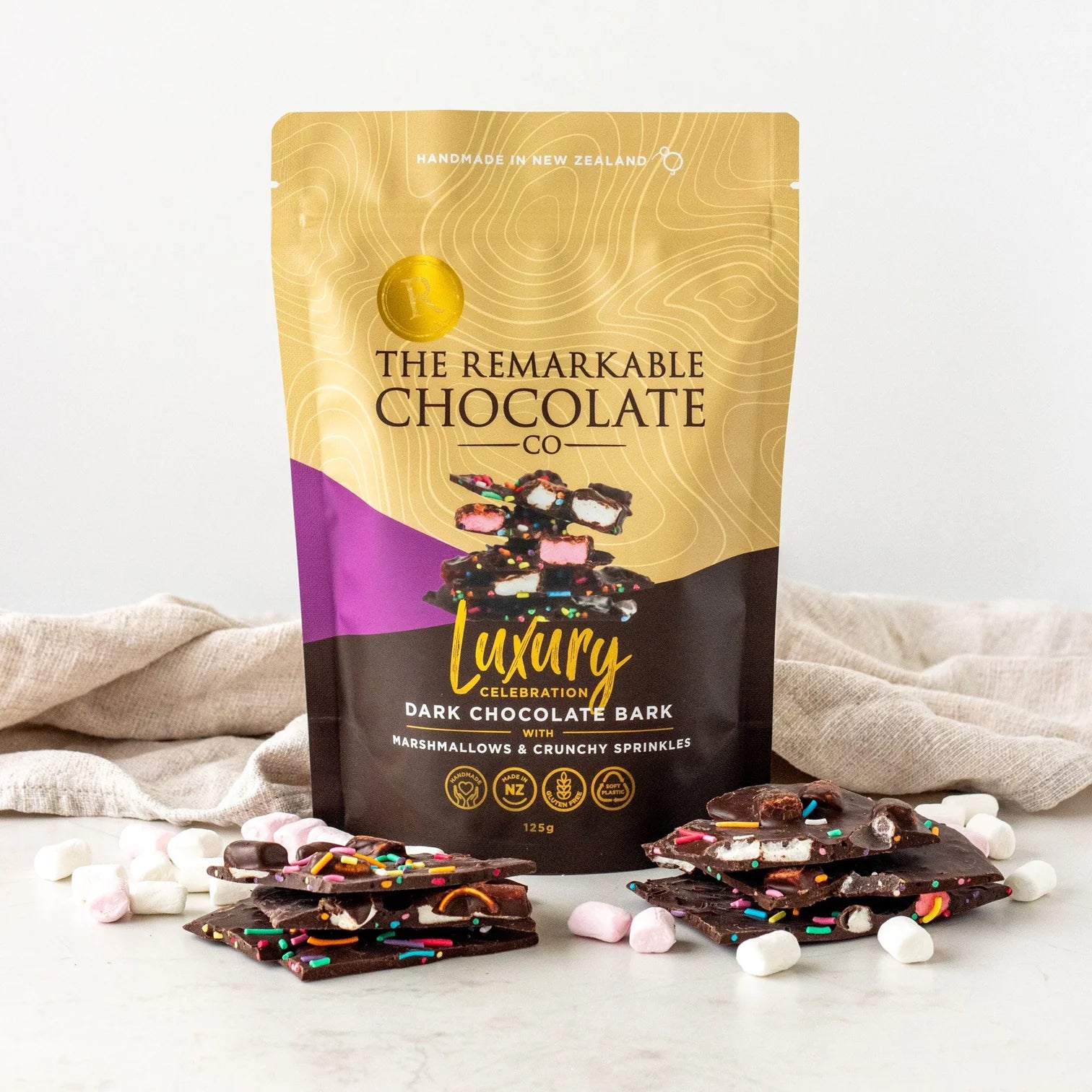Packaging of The Remarkable Chocolate Co. Luxury Dark Chocolate Bark mashmallow and crunchy sprinkles with some pieces displayed on a white surface.