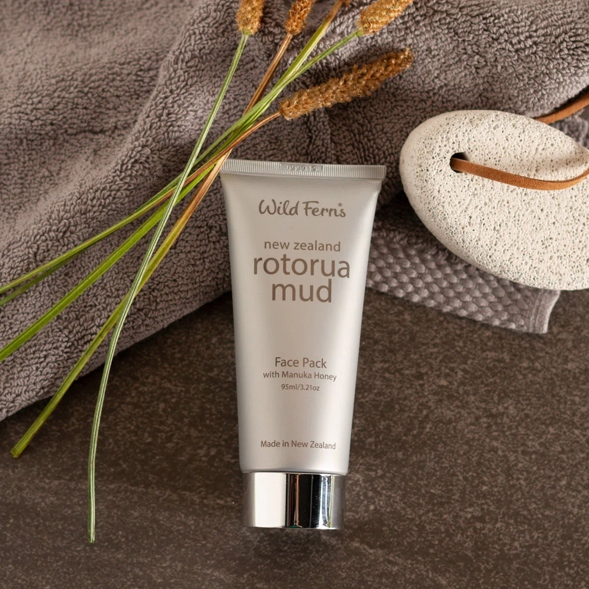 Tube of Wild Ferns Rotorua Mud Face Pack on a brown towel with plants and a loofah.