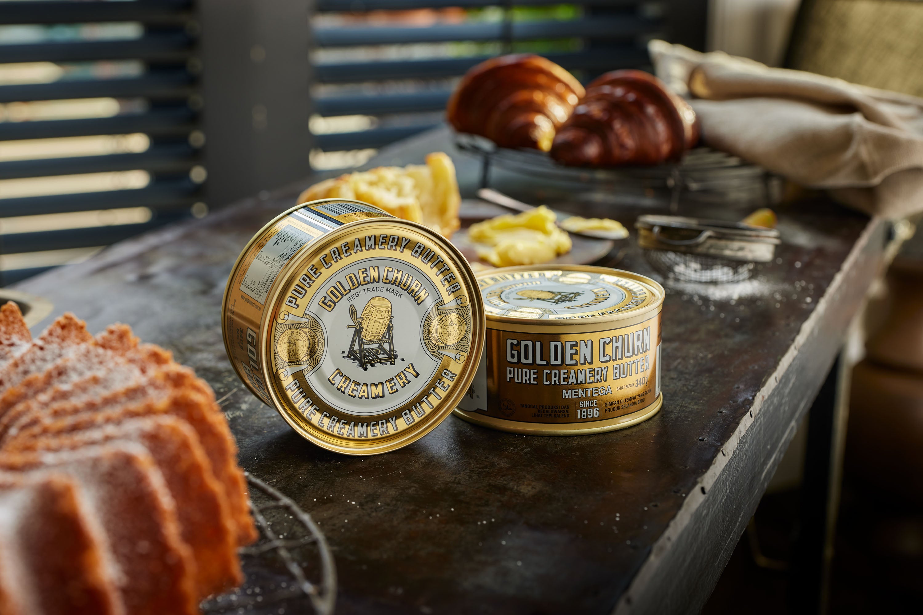 Two tins of Golden Churn Pure Creamy Butter on a rustic wooden surface with baked goods.