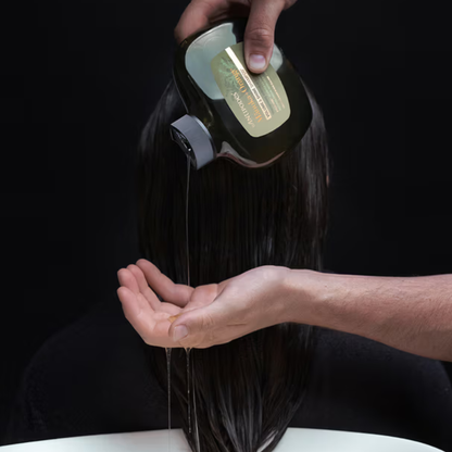 Person pouring liquid from a bottle  of Amtipodes Manuka Orange Shampoo onto their hair against a black background
