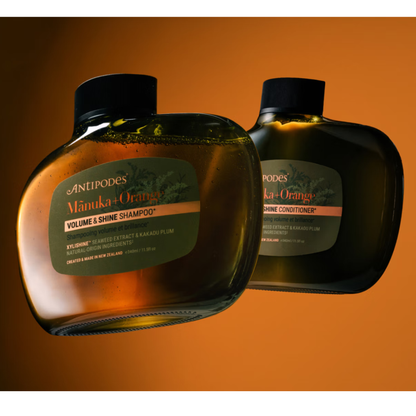 Two bottles of Antipodes Manuka + Orange Volume & Shine Shampoo and Conditioner on an orange background