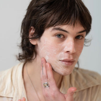 A close-up portrait of a person with white cream or soap applied to their cheek, touching their face with their hand. They are wearing a light beige collared shirt and a silver ring, set against a plain light gray background.