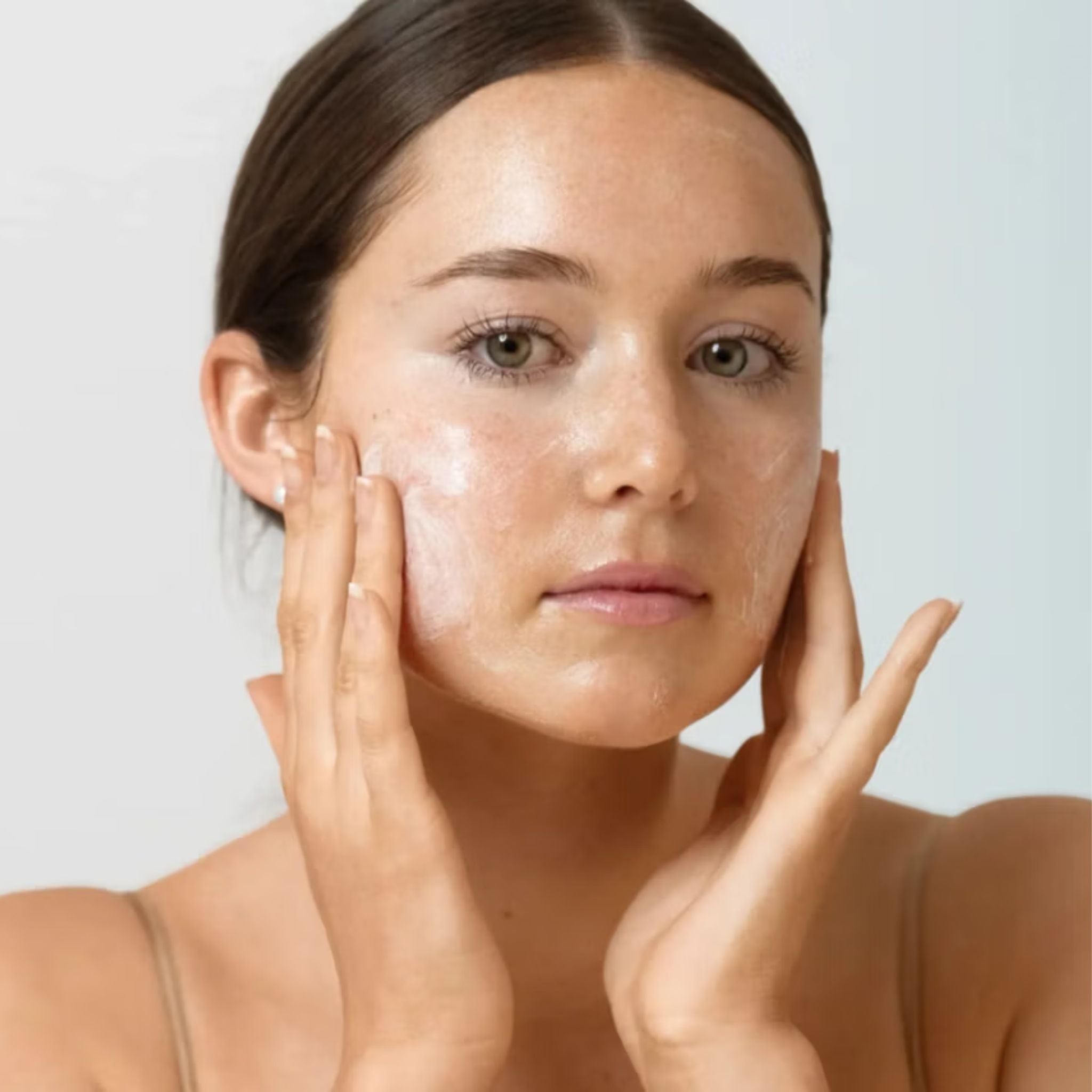 A close-up of a woman with clear, glowing skin touching her face with both hands against a light gray background.