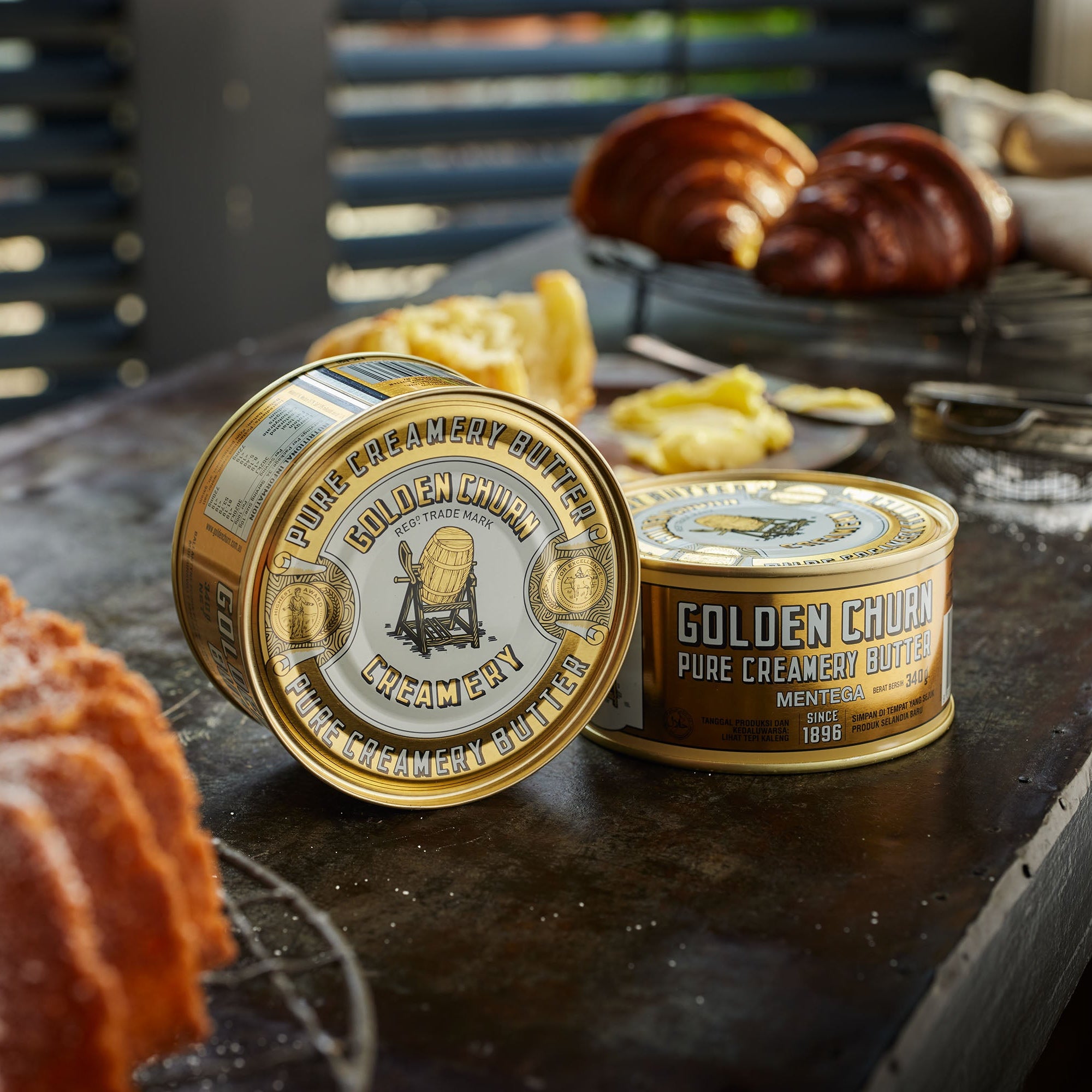 Two tins of Golden Churn Pure Creamy Butter on a rustic wooden surface with baked goods.
