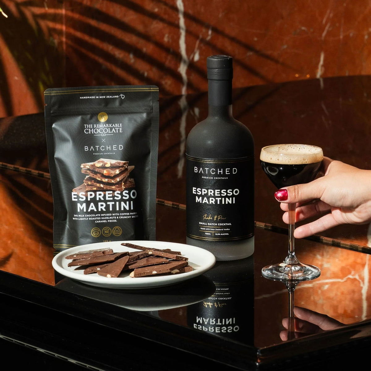 A bottle of The Brackmore Chocolate Espresso Martini and a bag of Batched Espresso Martini chocolate pieces are displayed on a dark reflective table next to a plate of chocolate shards and a hand holding a glass of espresso with foam.