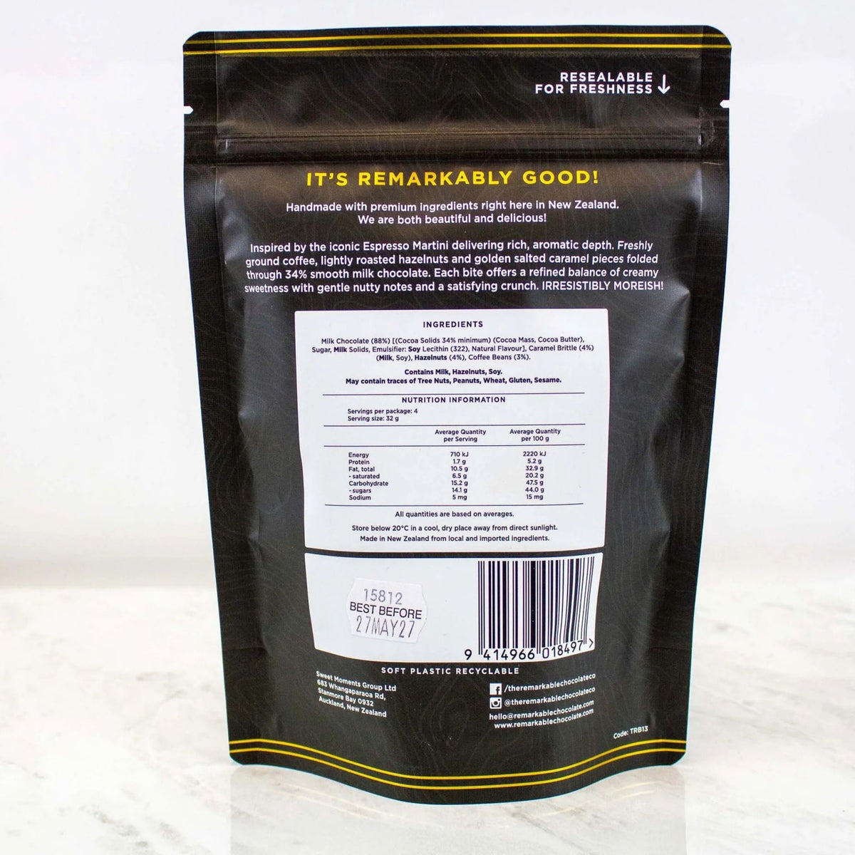 Back view of a resealable stand-up pouch of coffee on a light marble surface. The black pouch has yellow text and borders, with a large white ingredient panel in the center and a barcode near the bottom.