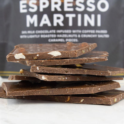 A stack of dark chocolate bark pieces with visible nuts and caramel pieces, set against a dark bag labeled 'ESPRESSO MARTINI'.