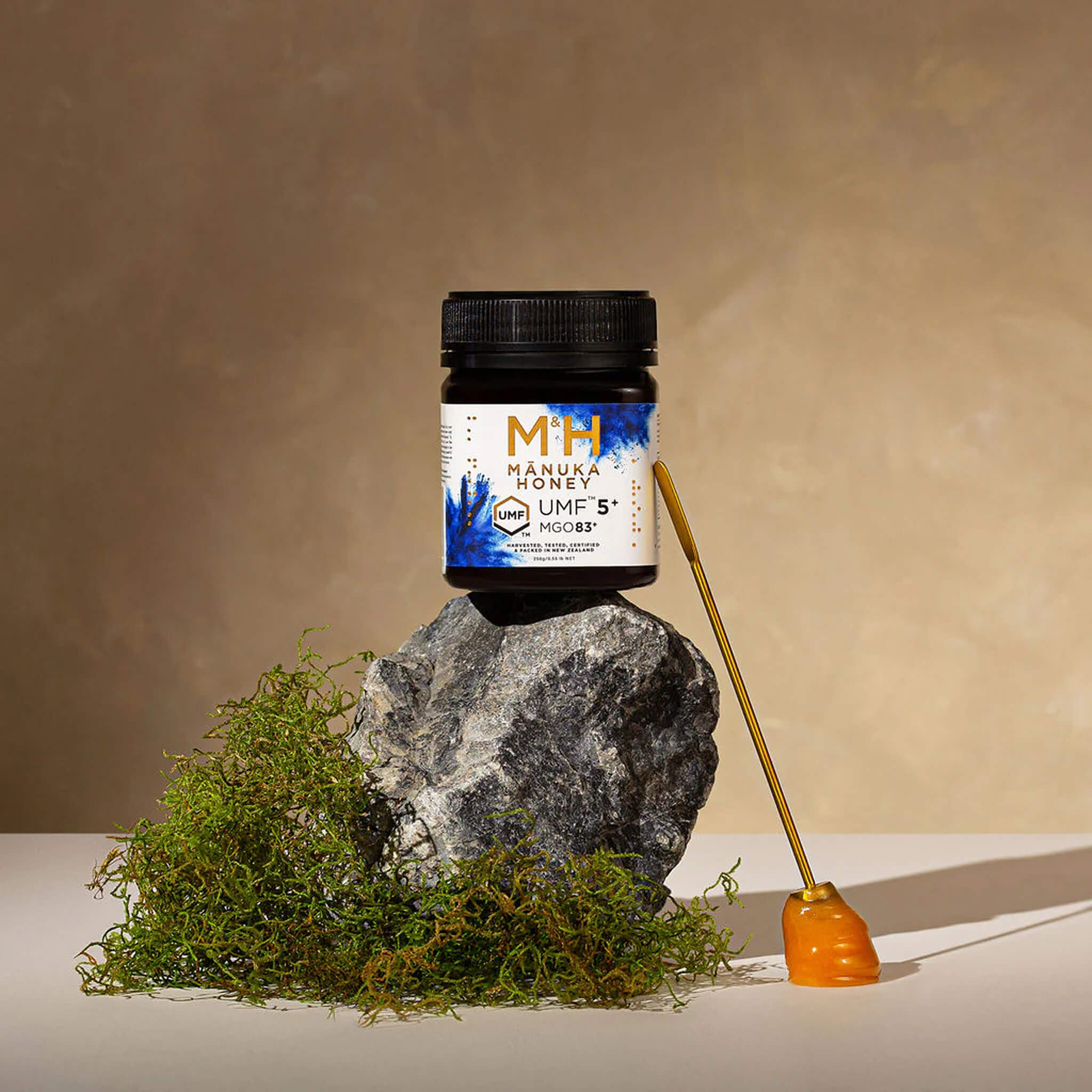 M&H Manuka Honey UMF5+ 250g jar with honey dripper on a stone and moss background