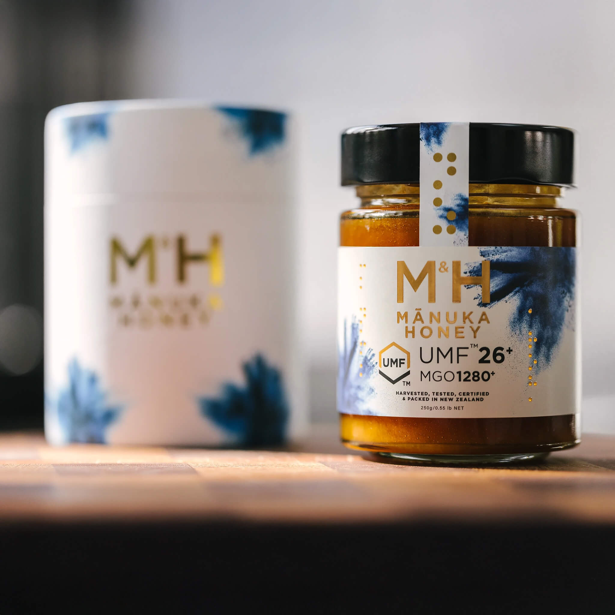 M&H Manuka Honey UMF 26+ jar with a branded mug on a wooden surface