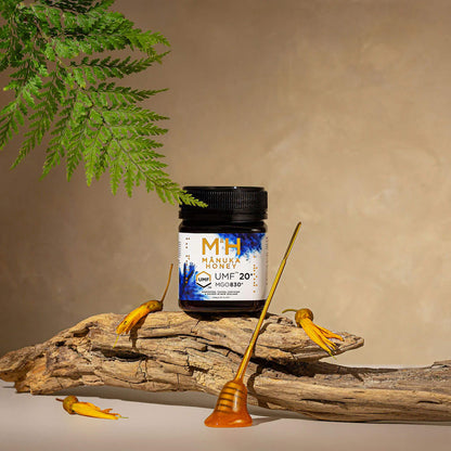 Manuka Health M·H UMF 20+ 250g jar on a wooden surface with honey sticks and fern leaves.