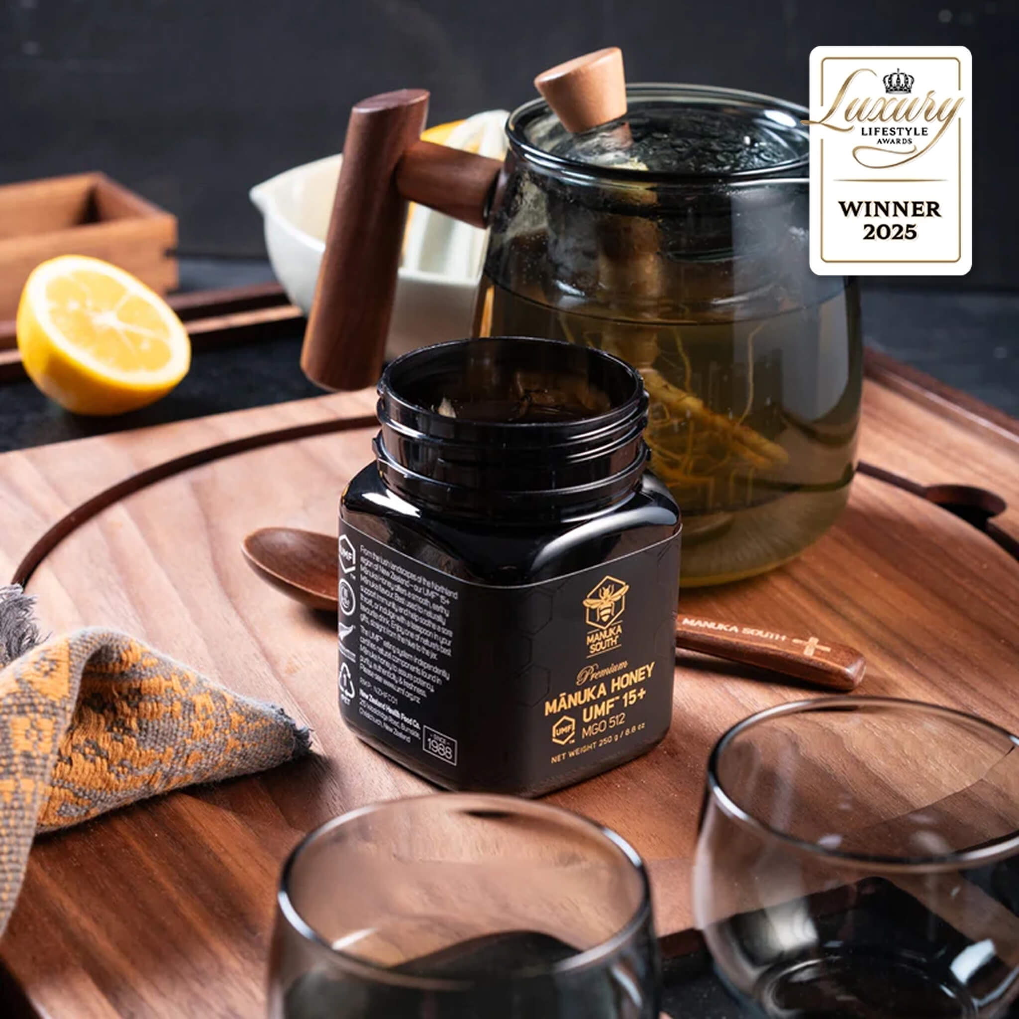 Manuka South Manuka honey jUMF 15+ 250g ar on a wooden table with glasses and lemon, featuring a 'Winner 2025' badge.