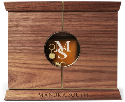 A rectangular wooden plaque or keepsake box with a warm medium-brown wood grain finish, featuring a circular brass emblem in the center with a white stylized 'MS' monogram and gold honeycomb details, and the words 'MANUKA SOUTH' engraved on the base. Manuka South Manuka Hosney UMF35+on a workshop bench