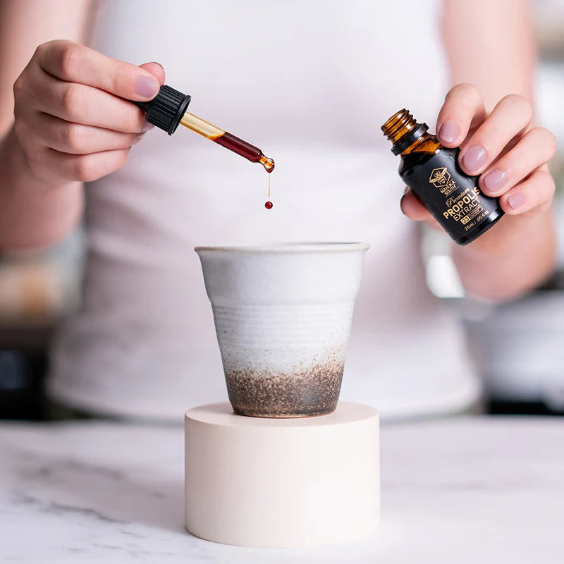 Person pouring propolis extract from a dropper bottle into a small cup on a white surface.