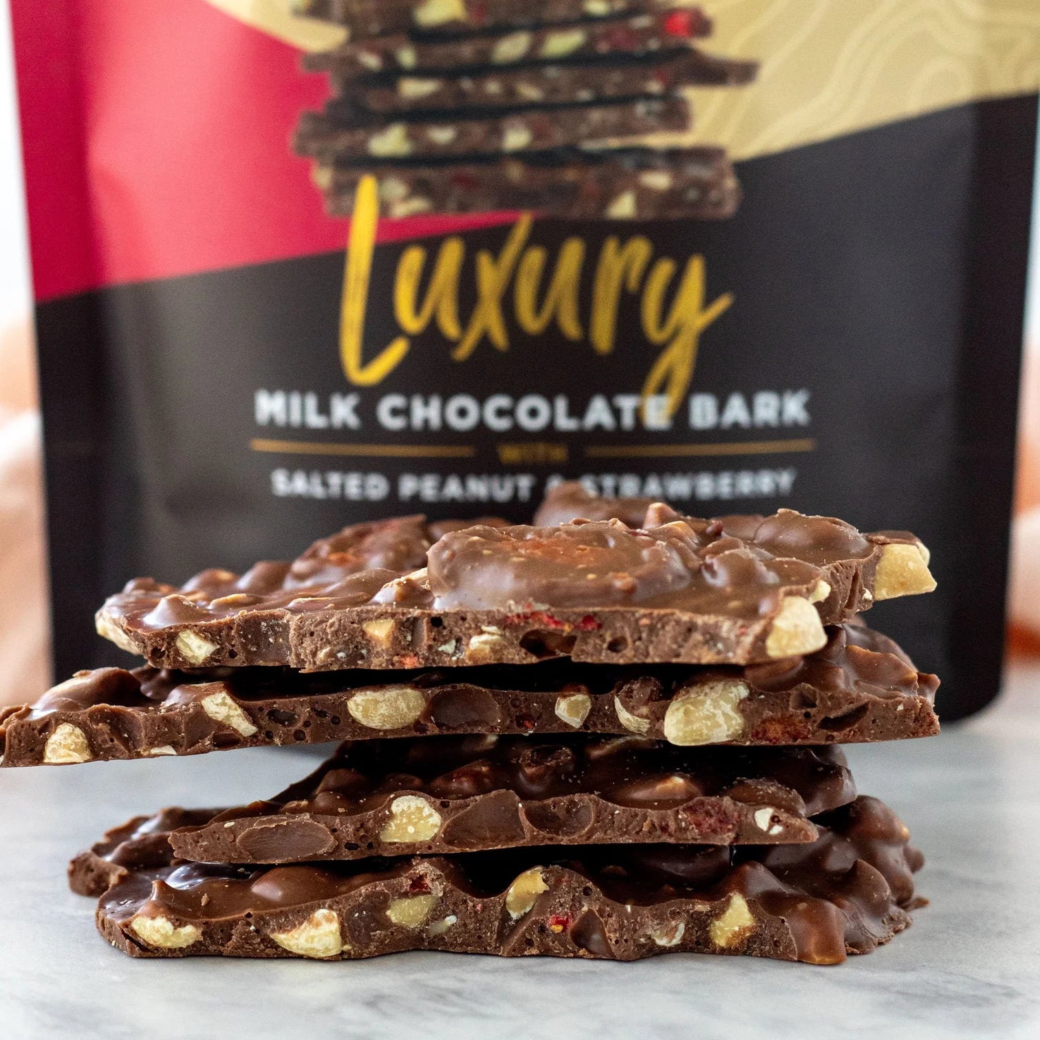 Stack of chocolate bark with 'Luxury Milk Chocolate Bark Salted Penut and Strawberry' packaging in the background.