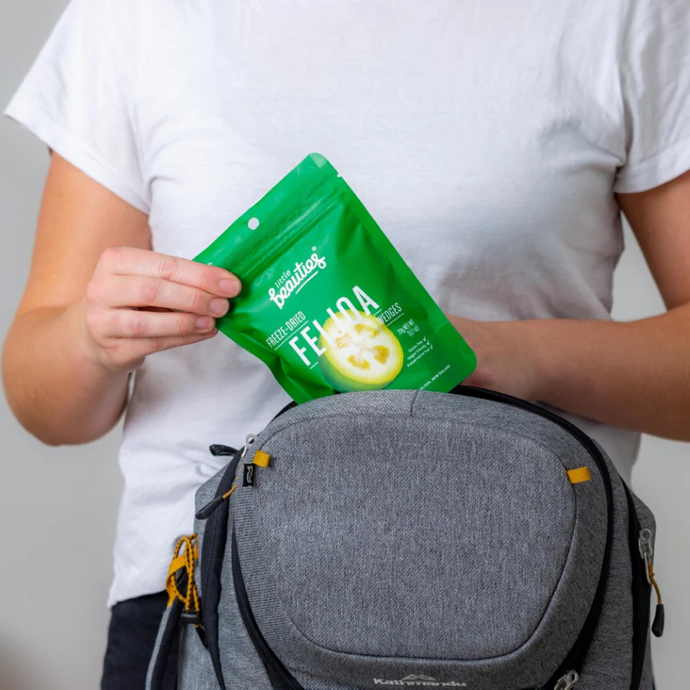 Person holding a green package of little beauties crunchy feijoa slices in front of a gray bag.