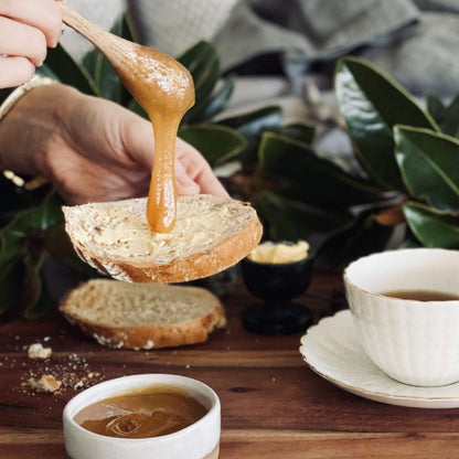 Person spreading a thick, manuka honey on a slice of bread with a wooden spoon, surrounded by greenery and a cup of tea.